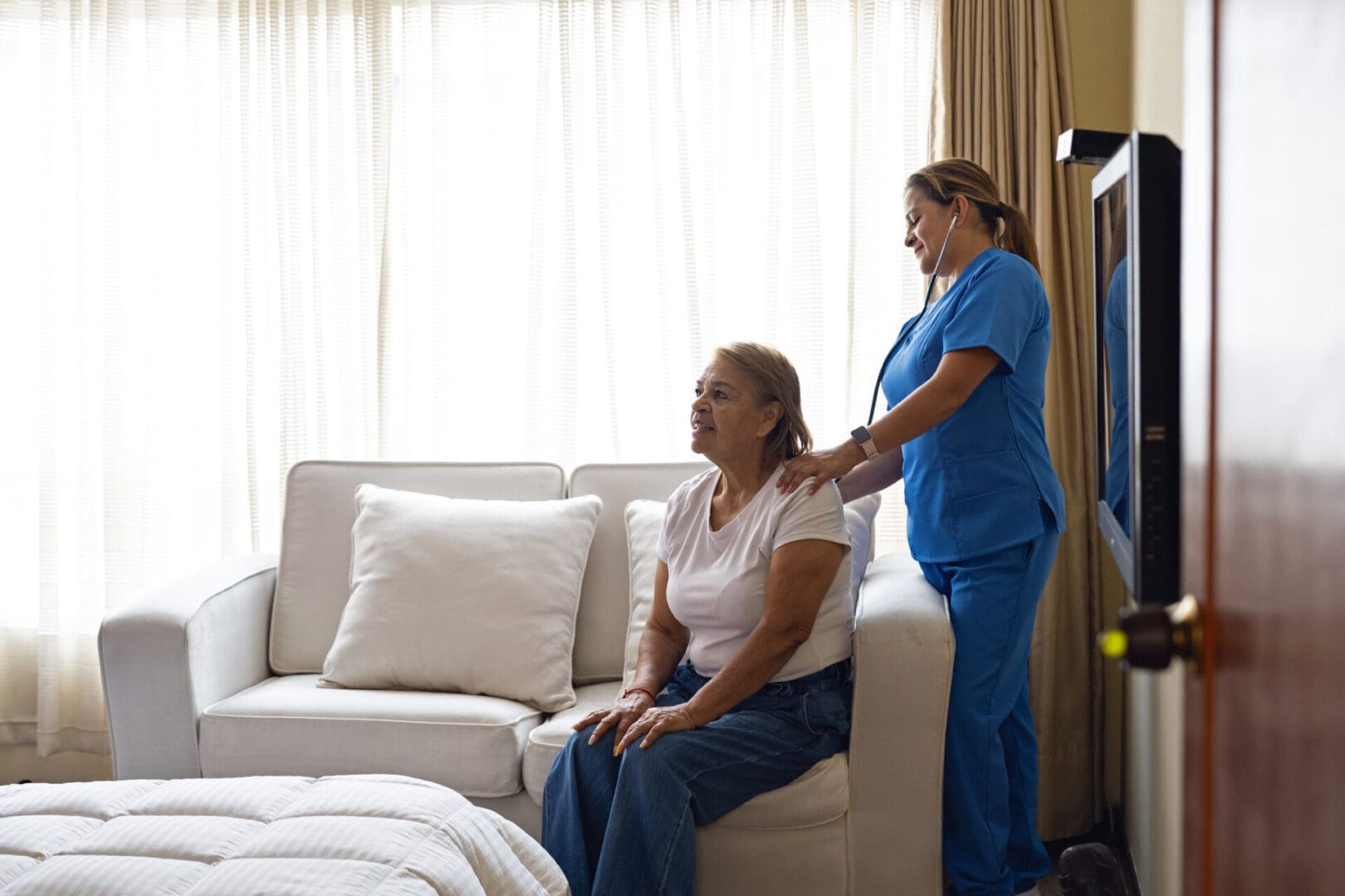 Nurse in blue scrubs uses a stethoscope to examine an elderly woman seated in her living room. Image illustrates personalized and preventive home healthcare for seniors with chronic conditions like diabetes or heart disease.
